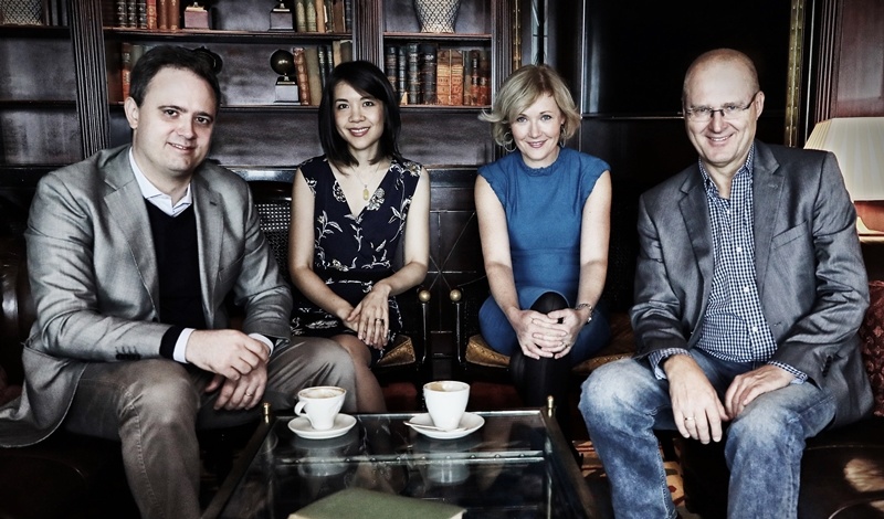 Vier Personen auf einer schwarzen Couch, im Hintergrund eine dunkle Bücherwand. In der Mitte, frontal zum Betrachter, sitzen zwei Frauen, rechts und links außen sitzt jeweils ein Mann. 