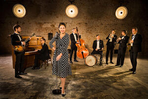 7 Musiker mit verschiedenen Instrumenten im Hintergrund. 
Eine Frau im gepunkteten Kleid mit Mikro im Vordergrund. 