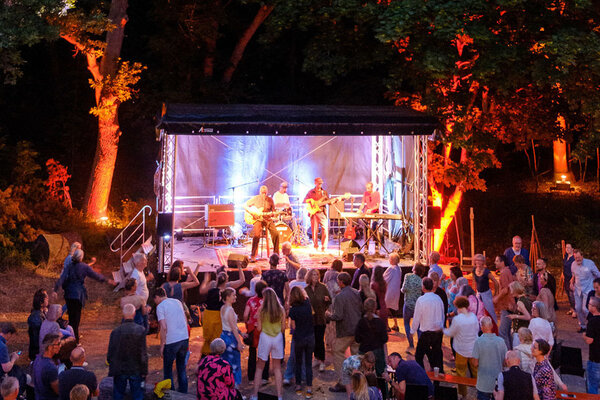 Bild vergr&ouml;&szlig;ern: Das Bild zeigt Menschen, die vor einer B&uuml;hne stehen, auf der Musiker ein Konzert geben.
