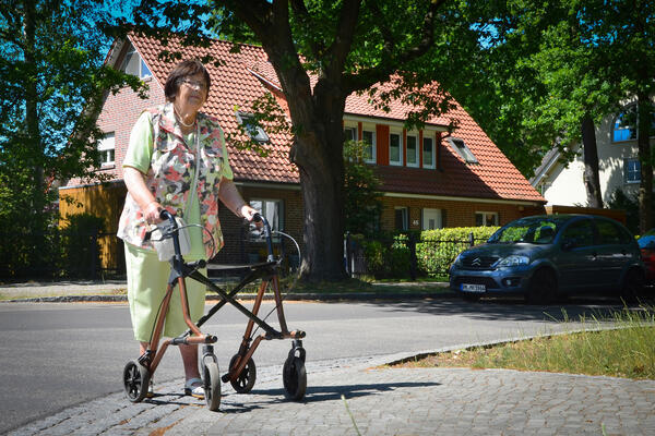 Bild vergrößern: Eine Dame mit Rollator auf der Straße im Hintergrund ein Haus