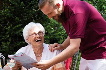 Bild vergrößern: Ein Pfleger reicht einer älteren Dame die Zeitung